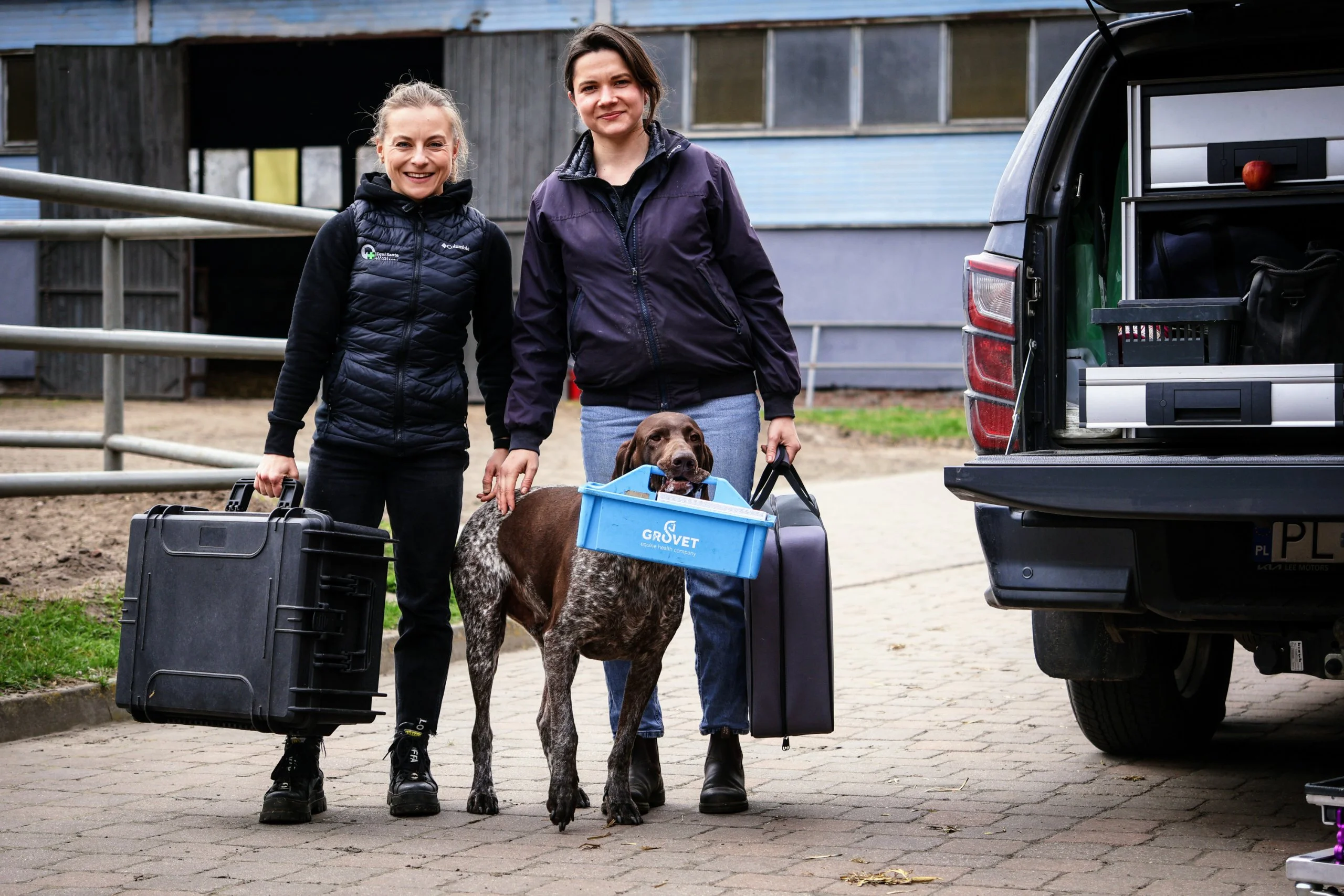 Lekarki Equi Sante idą od samochodu do stajni z walizkami sprzętu, obok biegnie pies